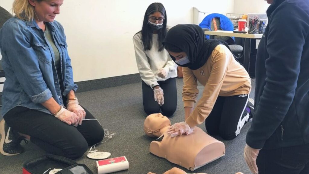 Four individuals are participating in a CPR training session. One person is practicing chest compressions on a CPR manikin, while others observe. An AED device is visible on the floor next to them.