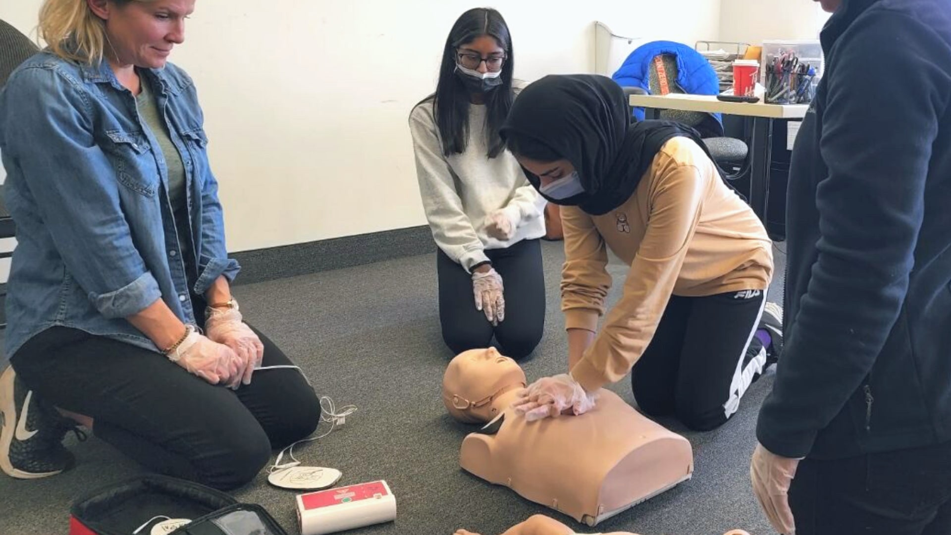 Four individuals are participating in a CPR training session. One person is practicing chest compressions on a CPR manikin, while others observe. An AED device is visible on the floor next to them.