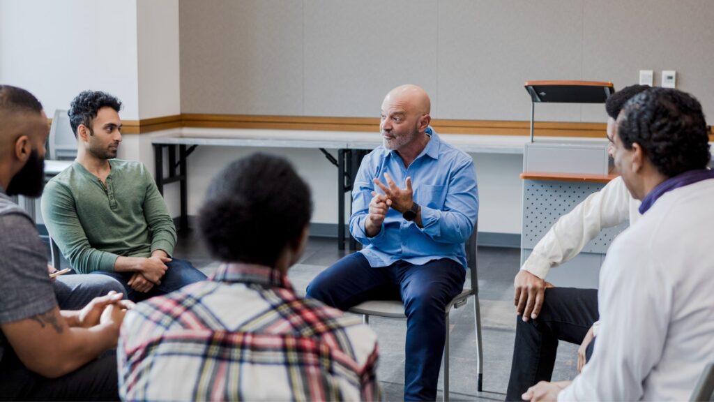 A diverse group of men sit in a circle in a room, engaged in a serious discussion. One man in a blue shirt gestures emphatically with his hands while speaking. They sit on chairs, with a lectern and a table visible in the background.