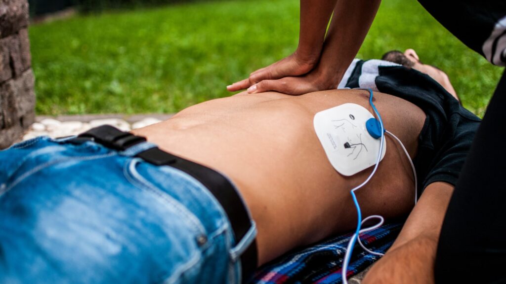 A person performs CPR on a man lying on a striped blanket outdoors. The man has a defibrillator pad on his bare chest. Another person assists nearby. Grass and a brick wall can be seen in the background.