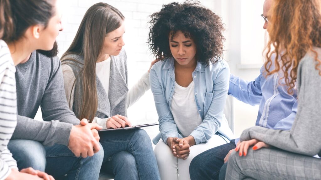A group of five people sit in a supportive circle. One person in the center looks upset, while others sit nearby offering comfort. They are in a bright room with natural light.