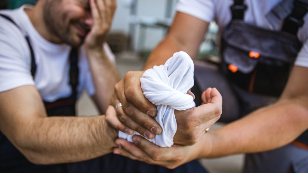 A man sits with his hand wrapped in a white cloth, appearing injured. Another person, wearing overalls, is helping by supporting the injured hand. The injured man is holding his head with his other hand. The scene suggests an accident or injury.