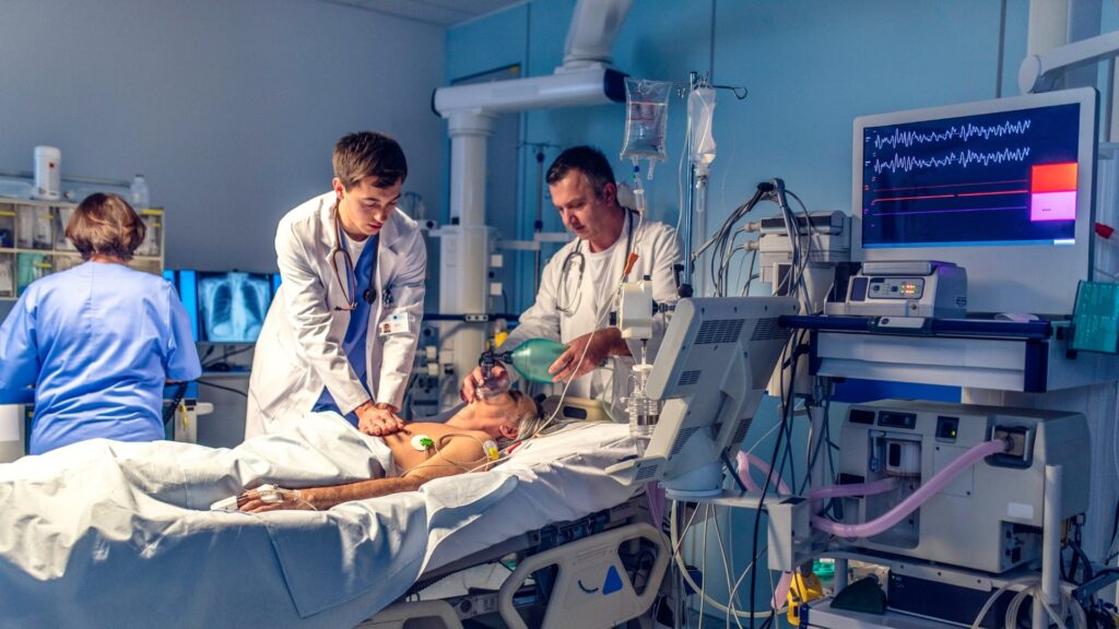 Two doctors and a nurse work on a patient in a hospital room. The patient lies in a bed connected to monitoring equipment, including a heart rate monitor showing vital signs. The doctors are focused on the patient, while the nurse checks equipment.