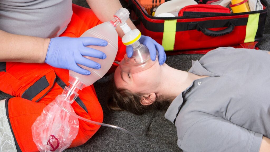 A person is lying on the ground receiving oxygen through a mask, administered by another person wearing gloves. A medical bag is visible in the background.