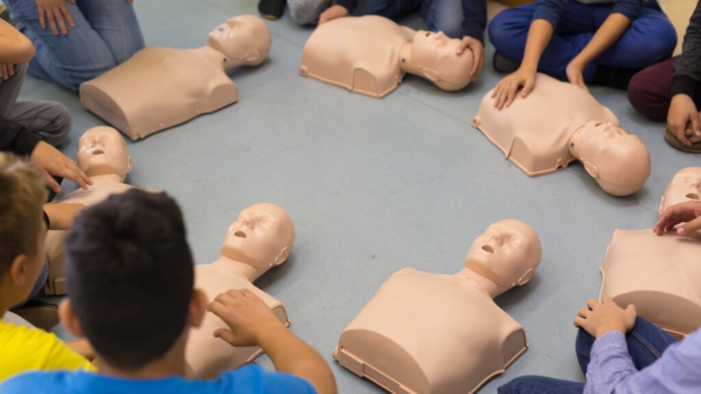 People sitting in a circle around CPR training manikins placed on the floor. The scene suggests a CPR training session, with participants preparing to practice on the manikins.