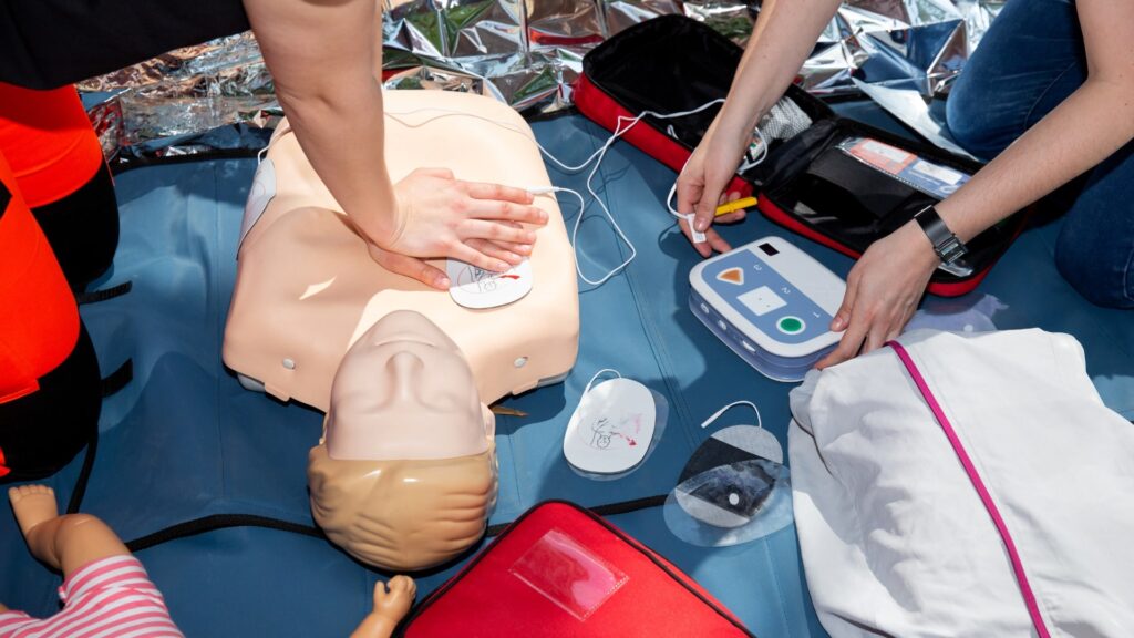 Two people practice CPR on a dummy. One person performs chest compressions, while the other prepares an AED device. Various emergency training equipment is visible, including a small mannequin and medical bag.