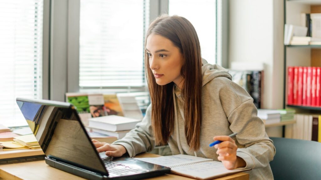 A woman with long brown hair is sitting at a desk, focused on her laptop. She's holding a blue pen and writing in a notebook. The room is bright, with books and papers nearby, and large windows in the background.