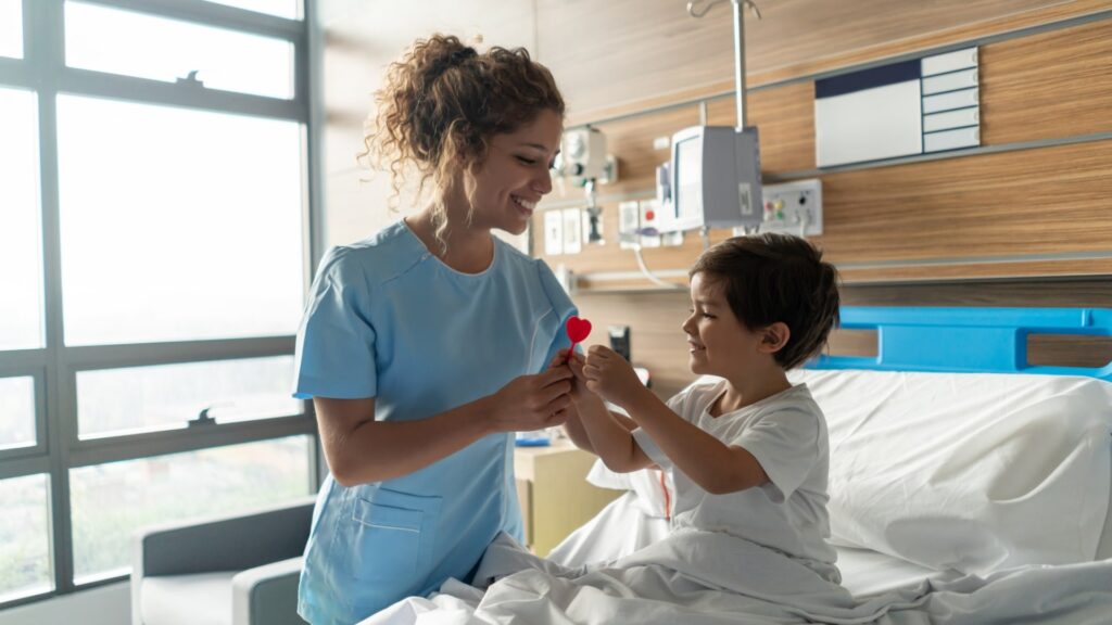 A nurse is smiling and handing a red heart object to a young boy sitting in a hospital bed. They appear to be in a hospital room with large windows, a wooden wall, and medical equipment in the background.