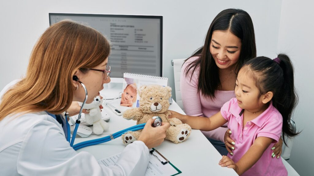 A doctor uses a stethoscope on a teddy bear while a child in a pink shirt and a woman smile at the scene. They are seated in a medical office, with a computer and calendar visible in the background.