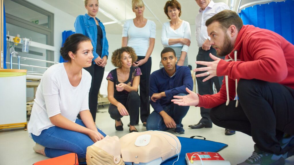 A group of people is gathered around a CPR training dummy on the floor. A man in a red hoodie is instructing, while others watch attentively. Some equipment is visible, indicating a first aid training session.