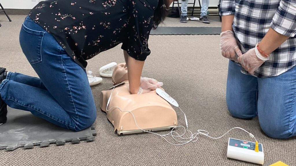 A person kneels on a mat performing CPR on a dummy, with another person kneeling nearby wearing gloves. An AED device is connected to the dummy. The setting appears to be a training session or class.