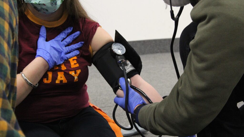 A person is having their blood pressure measured. They are wearing a maroon shirt with yellow text and have a hand placed on their chest. The healthcare worker uses a stethoscope and blood pressure cuff, wearing blue gloves.