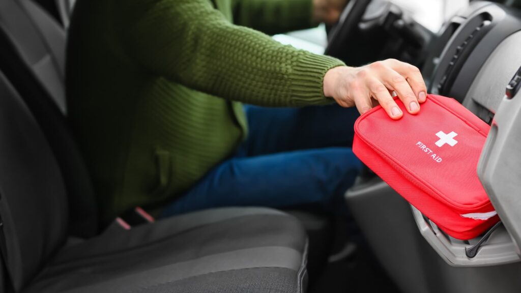 A person in a green sweater places a red first aid kit with a white cross into the glove compartment of a car. The person is seated in the driver's seat, and the interior of the car is visible.
