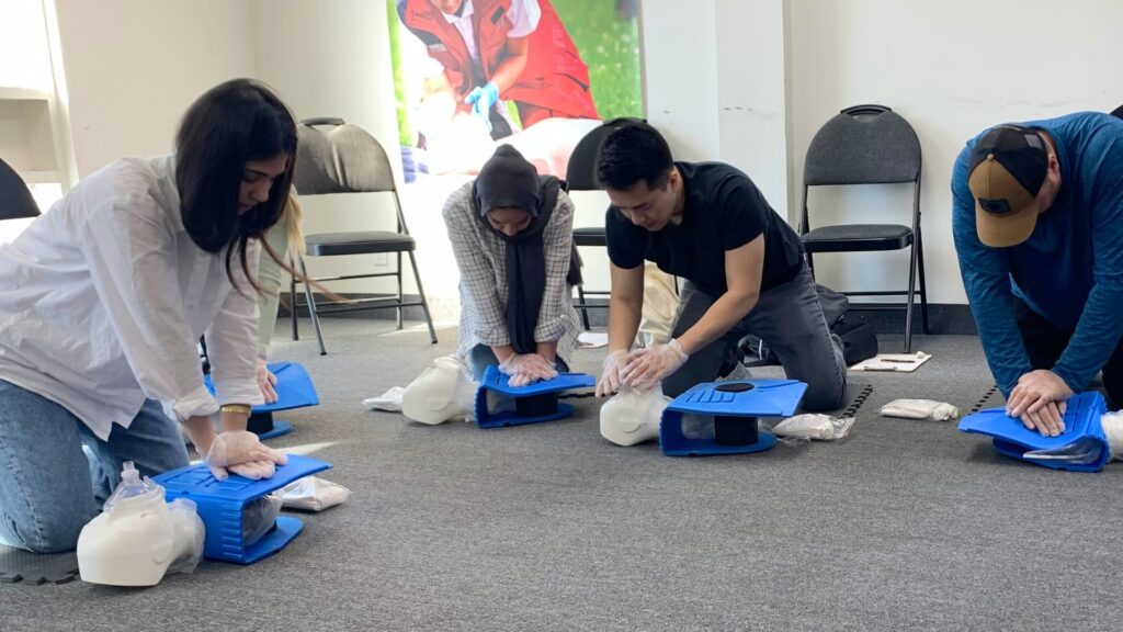 Four people practice CPR on training dummies in a classroom setting. They are kneeling on the floor, wearing gloves, and focusing on the activity. Empty chairs are in the background.