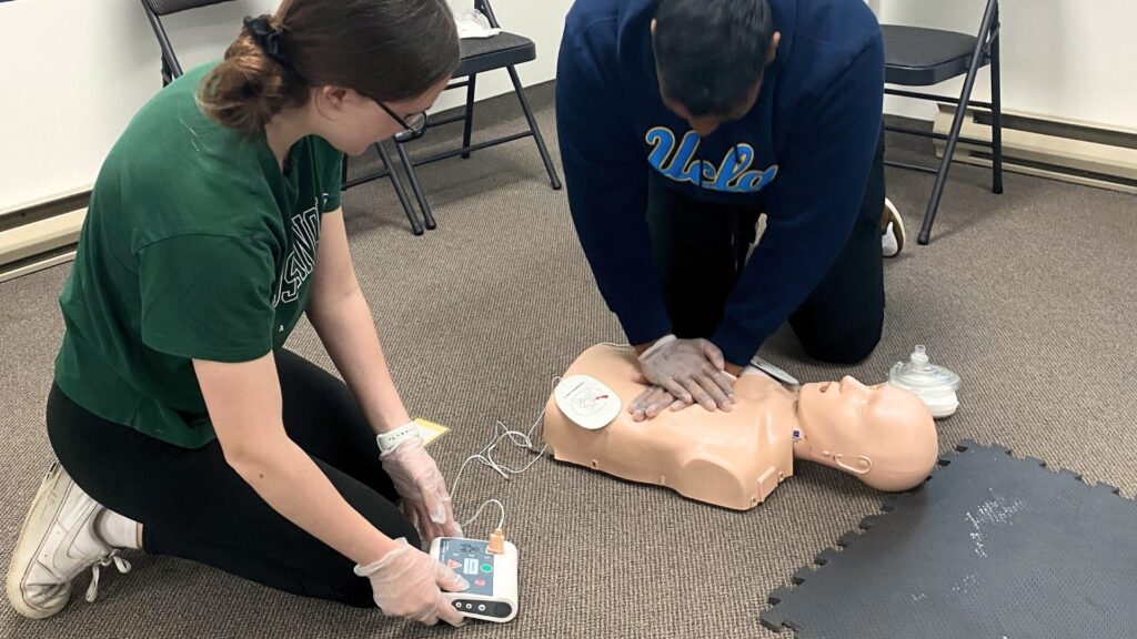 Two people are practicing CPR on a manikin. One person is performing chest compressions, while the other is operating a defibrillator. Both are wearing gloves, and there are chairs and a padded mat in the background.