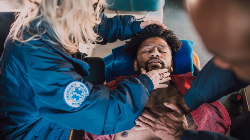 Emergency medical personnel attend to a man lying on a stretcher, checking his pulse and providing care. He is wearing a red jacket and appears to be unconscious. The scene is tense, showcasing urgent medical attention.