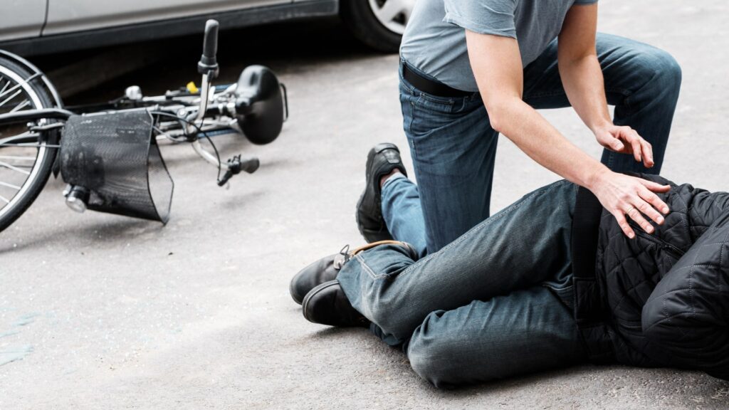 A person kneels on the ground assisting another individual lying down, with a bicycle fallen nearby. A car is parked in the background. Both are wearing casual attire, indicating a possible accident scene.