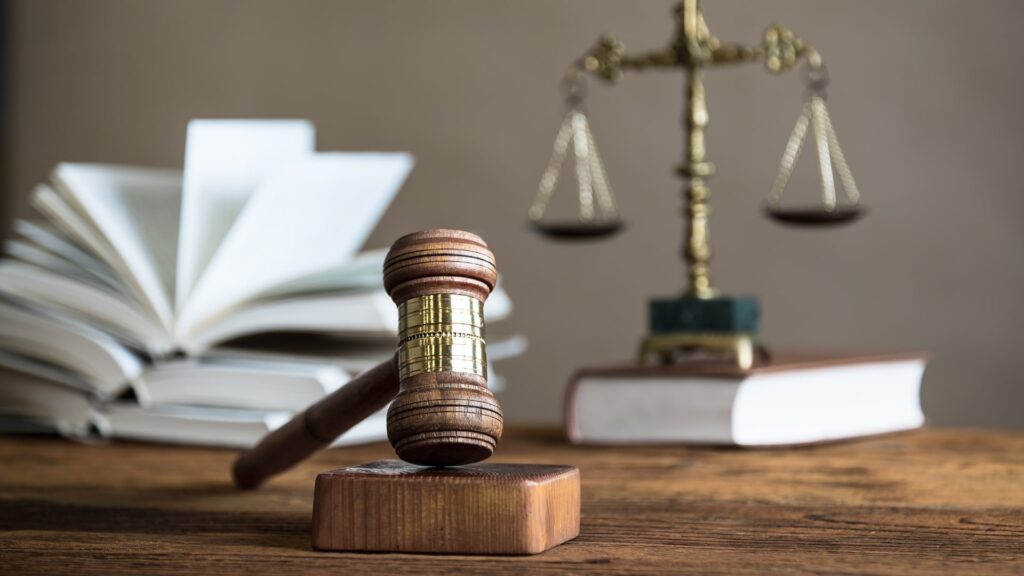 A wooden gavel rests on a sound block on a wooden table. In the background, there are open law books and a bronze balance scale. The setting suggests a legal or judicial theme.