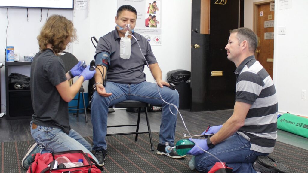 Two people kneel, wearing gloves, administering oxygen and taking a man's blood pressure. The man sits on a chair with an oxygen mask. The scene appears to be a first aid or medical training setting, with equipment bags nearby.