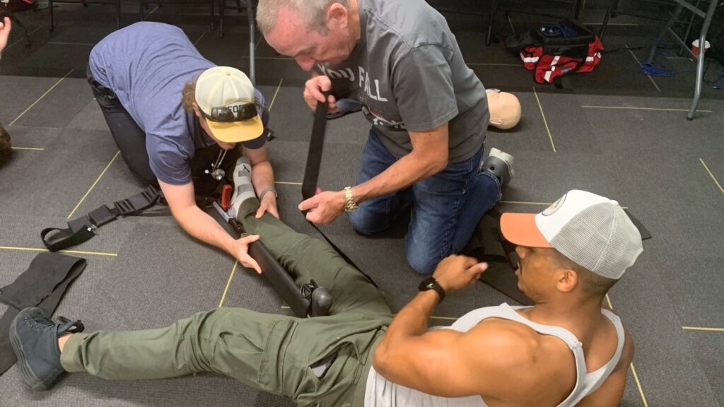 Three men practice applying a tourniquet on someone's leg in a training scenario. Two men kneel beside the person, adjusting the device, while the third lies on his back, appearing to assist. The setting is indoors on a gray carpeted floor.