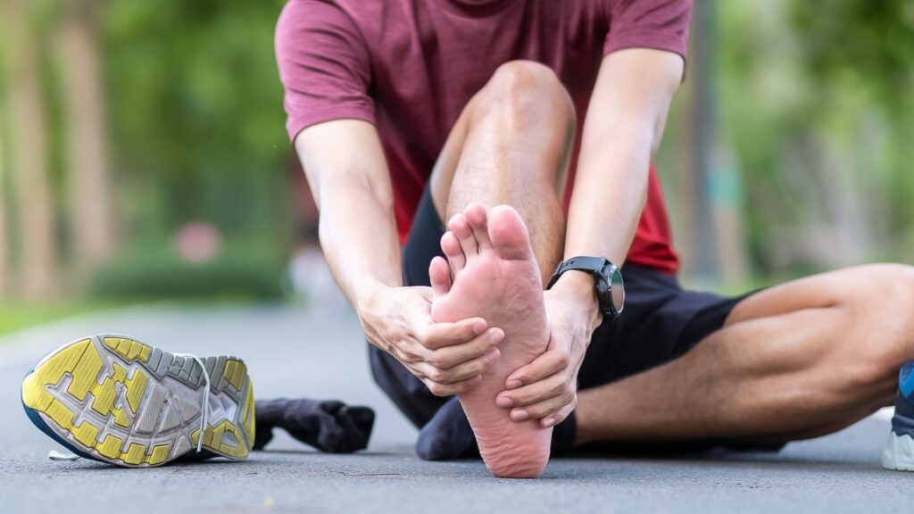 A person sitting on a pathway, holding their left foot with both hands, possibly stretching or examining it. Their right shoe and sock are off, placed to the left. They are wearing a red shirt and black shorts. The background is blurred greenery.