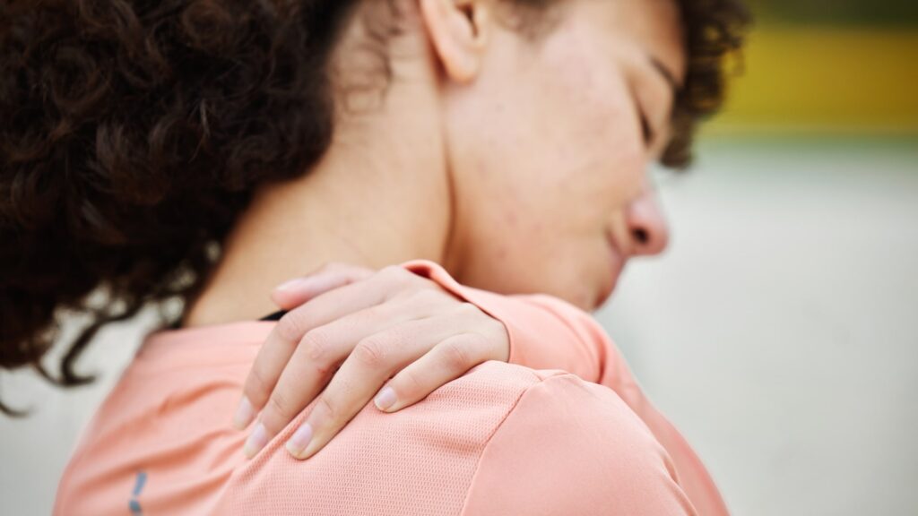 A person with curly hair wearing a pink top holds their shoulder, appearing to stretch or relieve discomfort. The image focuses on the hand and shoulder area, with a blurred background.