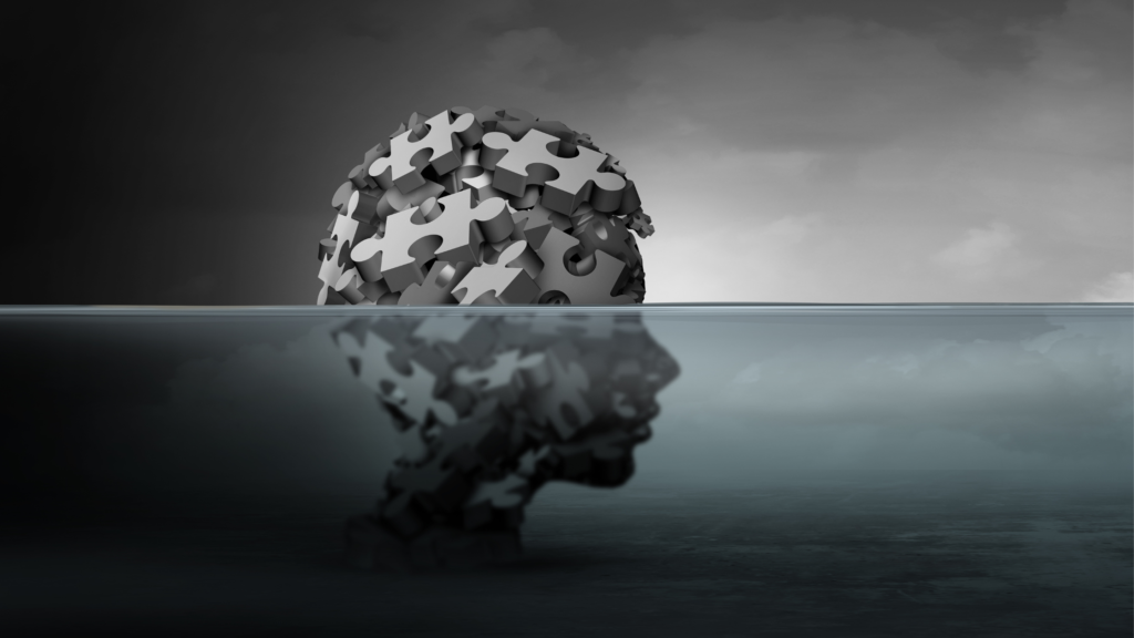 A silhouette of a human head composed of jigsaw puzzle pieces emerges from water, reflecting below the surface. The image is in grayscale, with a cloudy sky in the background, conveying themes of introspection and complexity.