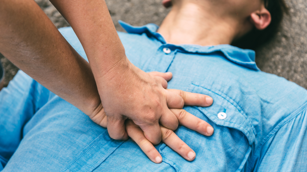 Person in a blue shirt lies on the ground while another person performs CPR by pressing hands on the chest. Hands focus on compressions, indicating a medical emergency or training scenario.