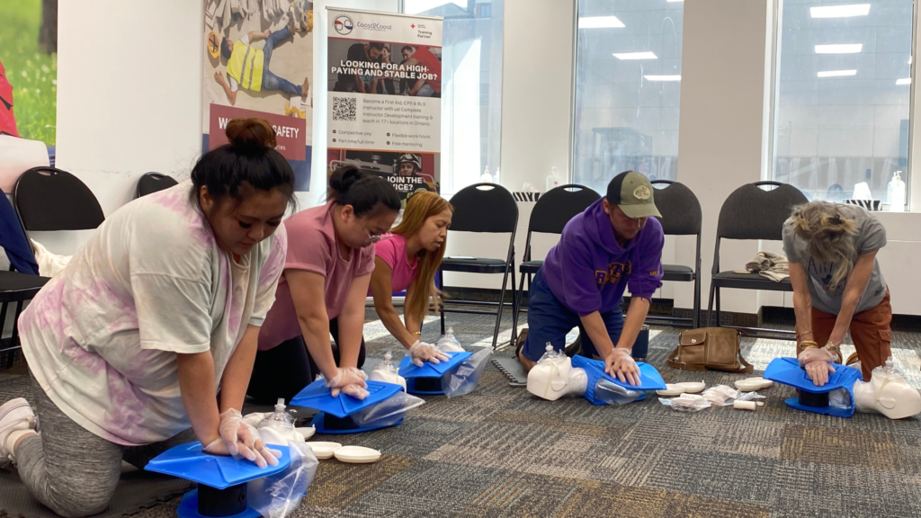Five people are kneeling on the floor in a training room, practicing CPR on blue training mannequins. They are focused on learning chest compressions. Chairs, posters, and CPR equipment are visible in the background.