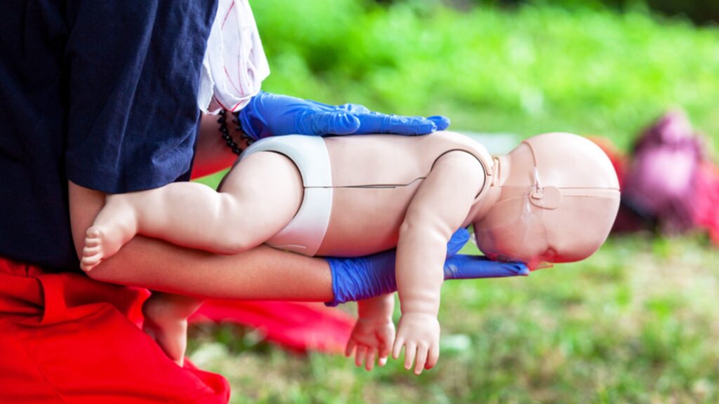 A person wearing blue gloves holds an infant CPR training mannequin, demonstrating a first aid technique for choking. The background is blurred greenery, indicating an outdoor setting.