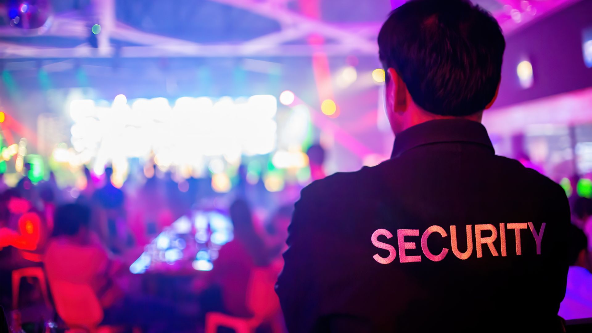 A security guard stands watch as a diverse group of people socialize under vibrant disco lights in a lively party atmosphere.