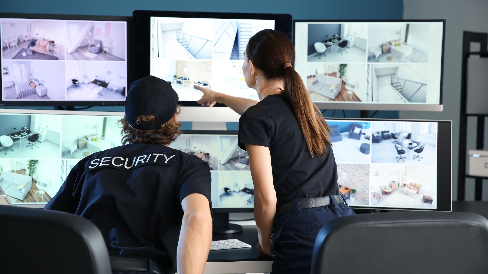 A female and male security personnel collaborate in a security office, analyzing surveillance footage displayed across multiple monitoring screens; one gestures toward a specific screen segment while the other observes closely