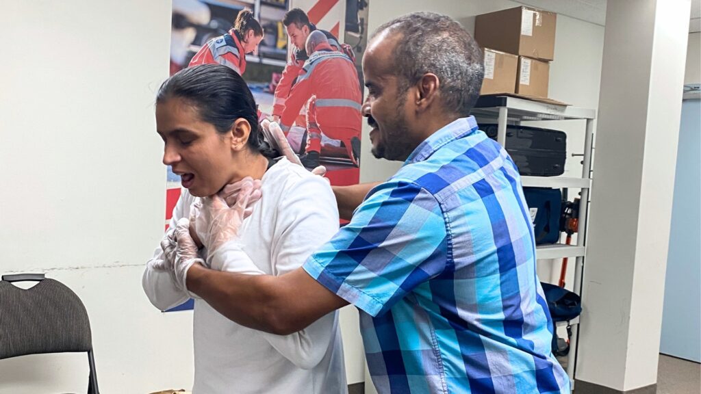 A man in a blue checkered shirt performs the Heimlich maneuver on a woman in a white sweater who appears to be choking. They are indoors, with chairs and boxes in the background.