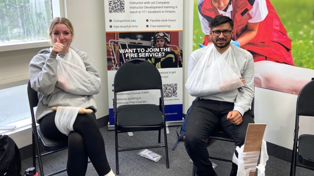 Two people sitting on chairs with their arms in slings, smiling. A poster behind them reads, "Want to Join the Fire Service?" indicating they might be in a first aid or emergency response training setting.