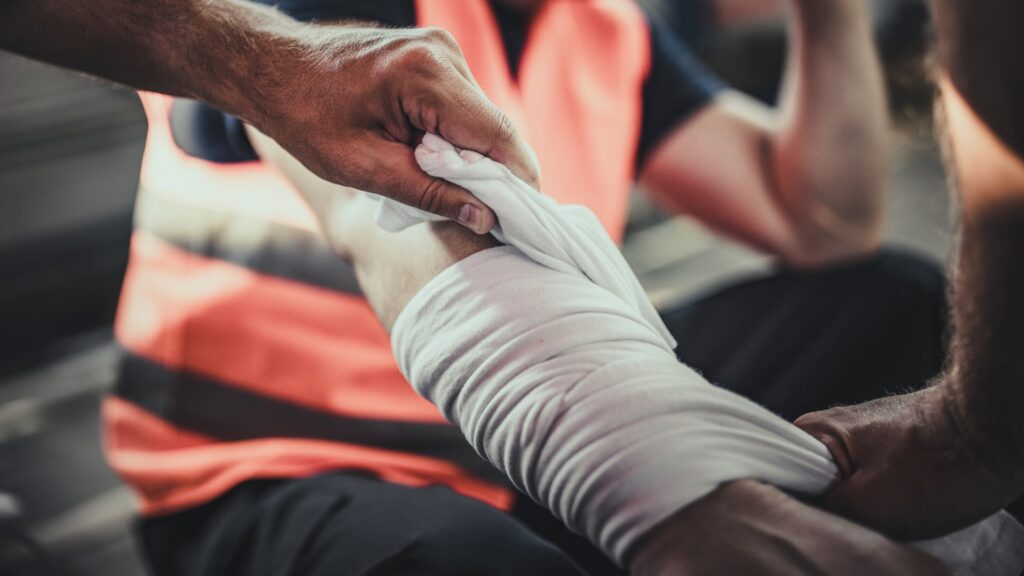 A person in an orange safety vest is wrapping a bandage around another person's arm. The scene suggests first aid is being administered, possibly in an emergency or outdoor setting. There is a focus on hands and the bandaging process.