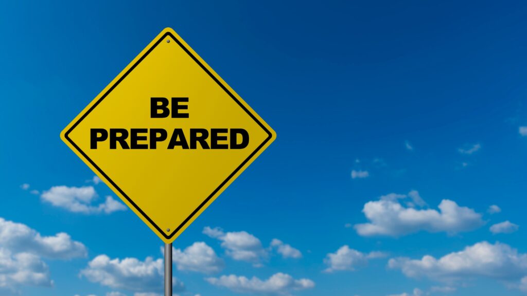 A yellow diamond-shaped road sign with the words "BE PREPARED" stands against a blue sky with scattered white clouds.