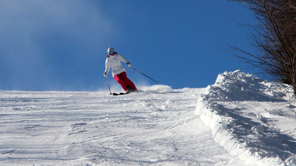 A skier wearing a white jacket and red pants descends a snowy slope under a clear blue sky. The skier is making a turn, kicking up powder as they go. Sparse trees are visible on the right side of the image.