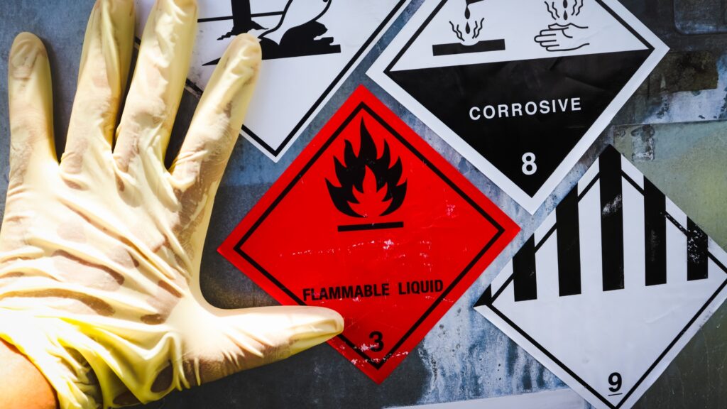 A gloved hand next to hazard symbols on a wall, including a red flammable liquid sign with a flame icon, a corrosive sign depicting liquid damaging a surface, and a black-and-white striped warning.