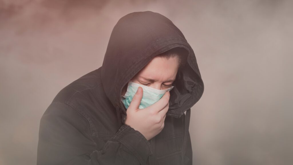 A person in a hooded jacket wearing a face mask appears to be coughing or covering their mouth with one hand amidst a smoky or foggy background.