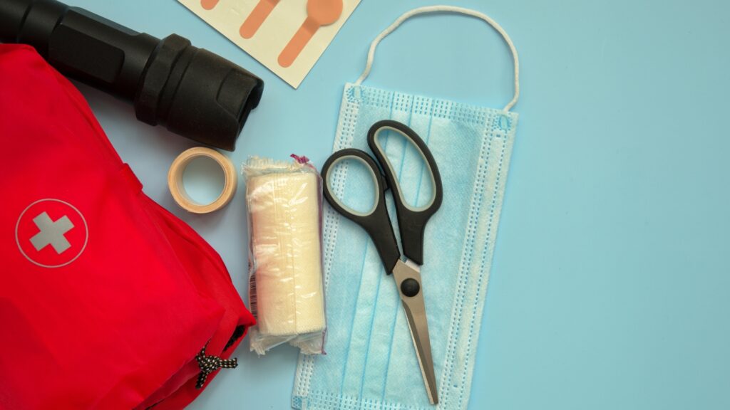 A flat lay image showing a red first aid kit, flashlight, adhesive bandage, roll of medical tape, wrapped bandage, pair of scissors, and a blue disposable mask on a light blue background.