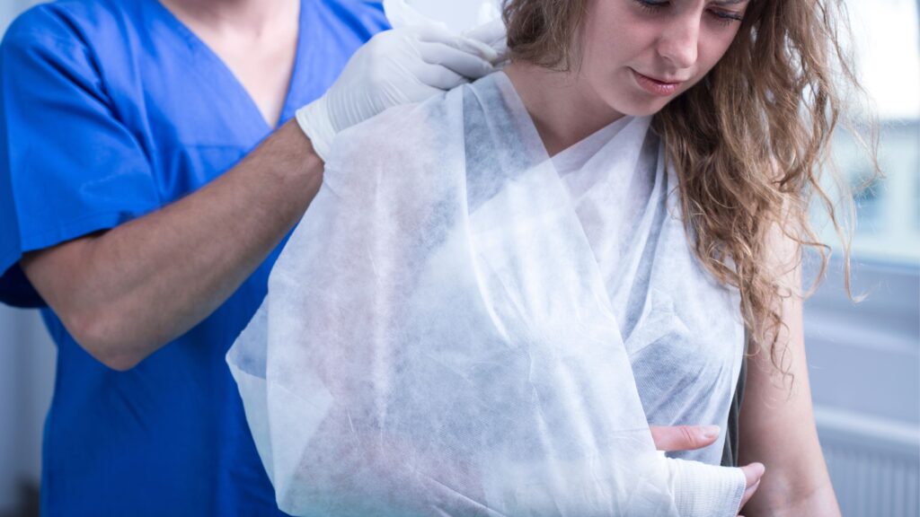A person in a blue medical uniform is assisting a woman with her arm in a cast and sling. The woman, with curly hair, looks down. They are indoors, in what appears to be a medical setting.
