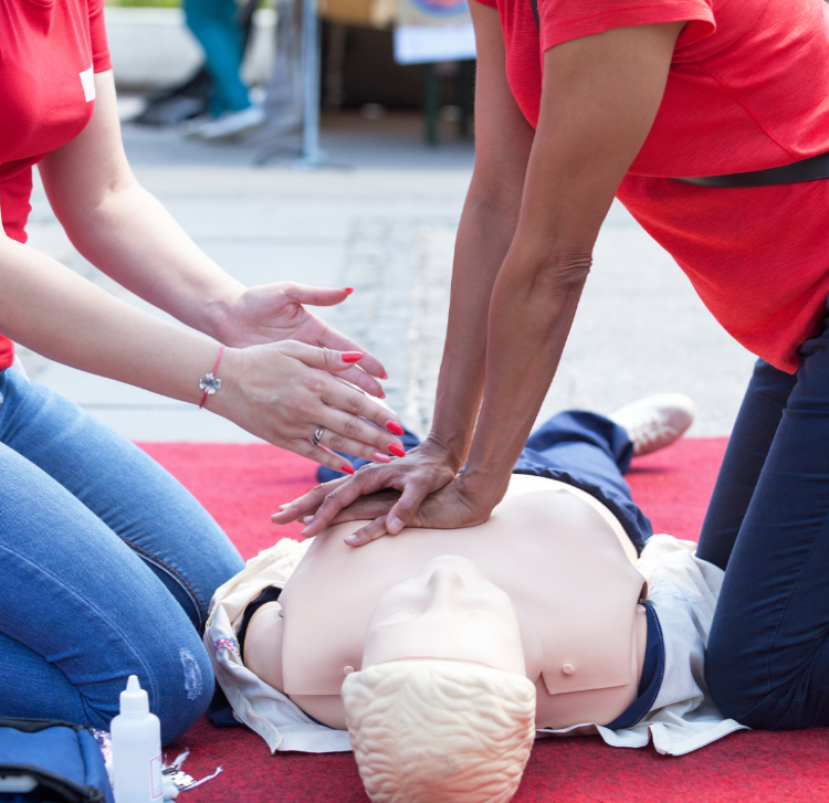 Two people practice CPR on a mannequin outdoors. One person performs chest compressions while the other watches closely. Both wear red shirts, and the scene is set on a red carpet in an open space.