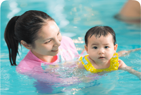 A woman in a pink shirt is holding a baby wearing a yellow floaty in a swimming pool. The woman is smiling at the baby, who looks toward the camera. The water is clear and blue.