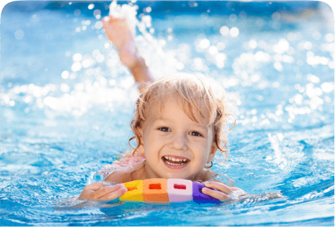 A young child with blonde hair happily swims in a pool, holding a colorful float. The water is clear and sparkling in the sunlight, creating a joyful and refreshing scene.