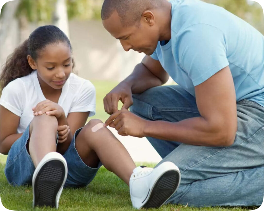 A man in a light blue t-shirt is kneeling and placing a bandage on a young girl's knee. The girl, wearing a white t-shirt and denim shorts, sits on the grass, watching attentively. Both appear calm and focused.