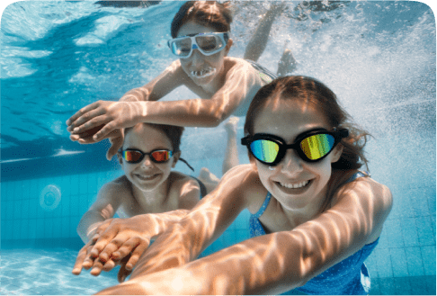 Three children swim underwater in a pool, wearing goggles. They smile and reach towards the camera, creating a joyful and playful scene. Light reflections ripple on the water, enhancing the bright and cheerful atmosphere.