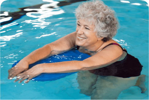 Elderly woman smiling and holding a blue kickboard while swimming in a pool. She appears relaxed and is wearing a dark swimsuit. The water around her is clear and blue.