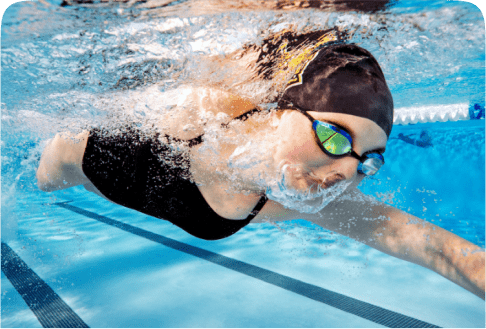 A swimmer wearing a black swimsuit and swim cap is gliding through clear blue water. They are wearing goggles and appear focused, with bubbles around their face. The pool's lane markings are visible below.