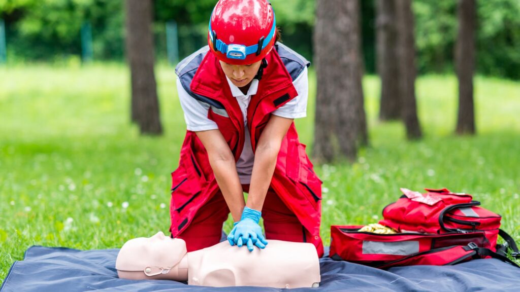Person in a red uniform and helmet performs CPR on a practice mannequin outdoors on a mat amid a grassy area with trees. A red emergency bag is beside them.