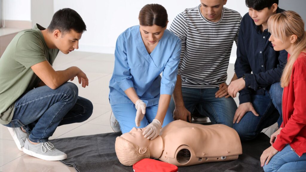 A group of five people attentively watch a medical professional in blue scrubs demonstrating CPR on a training mannequin. They are gathered around the mannequin, kneeling on the floor, with a red bag nearby.